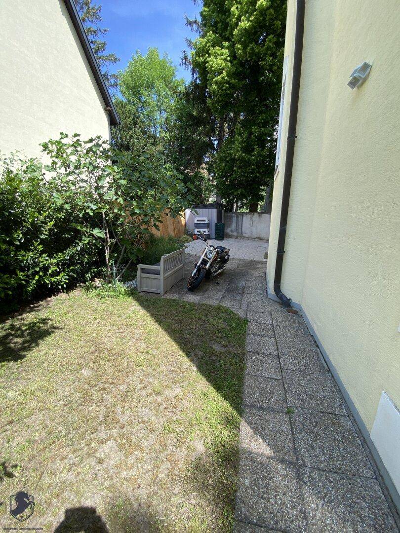 Seitlicher Gartenbereich mit Rasenfläche, gepflastertem Weg und einer Bank im Freien.