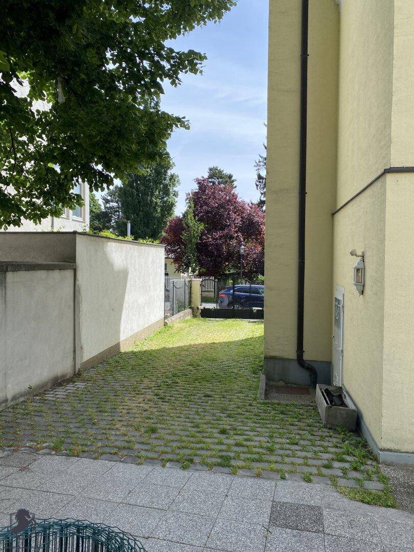 Seitlicher Gartenweg mit Gras zwischen Pflastersteinen und Blick auf weitere Bäume.