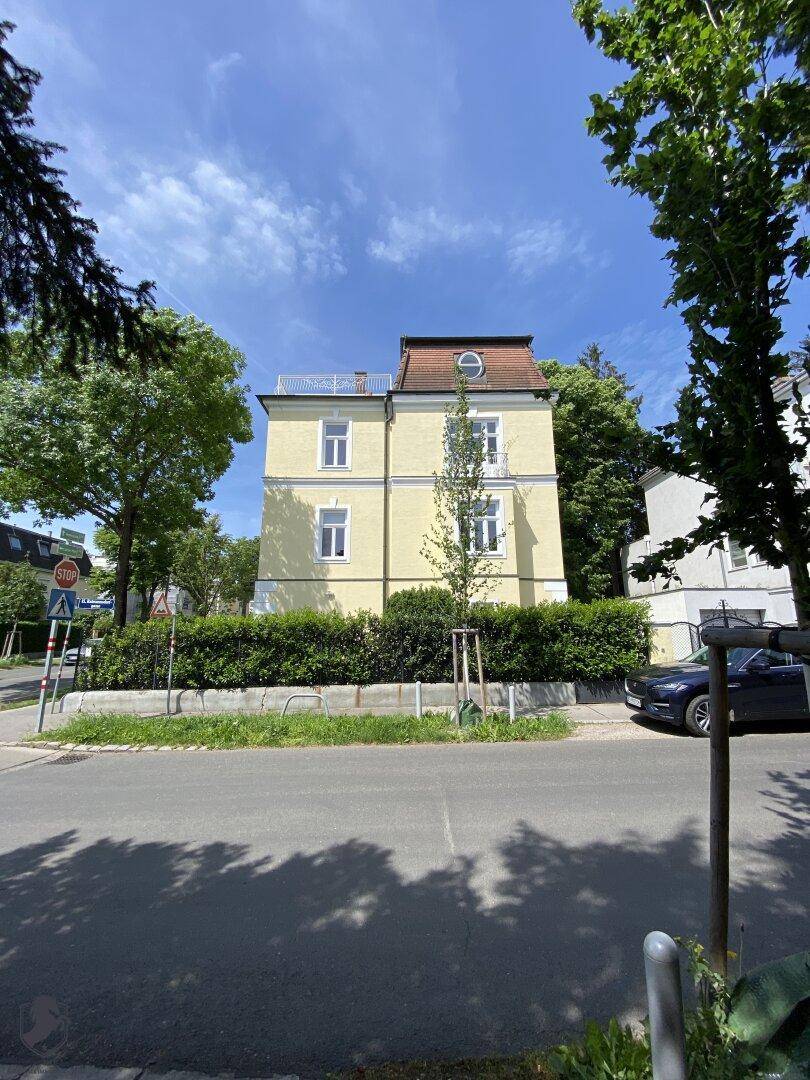 Gelbe Villa von der Straße aus gesehen mit Mansarddach und umliegenden Bäumen.