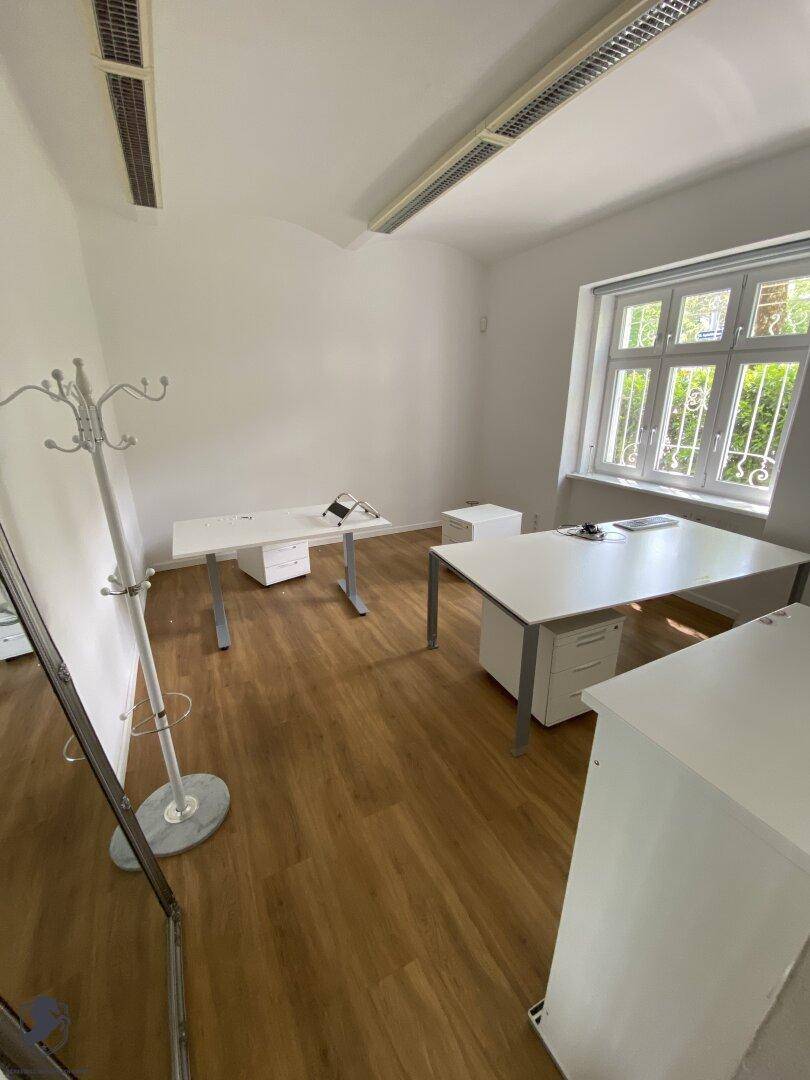Helles Büro mit Laminatboden, zwei Schreibtischen und großem Fenster mit Grünblick.