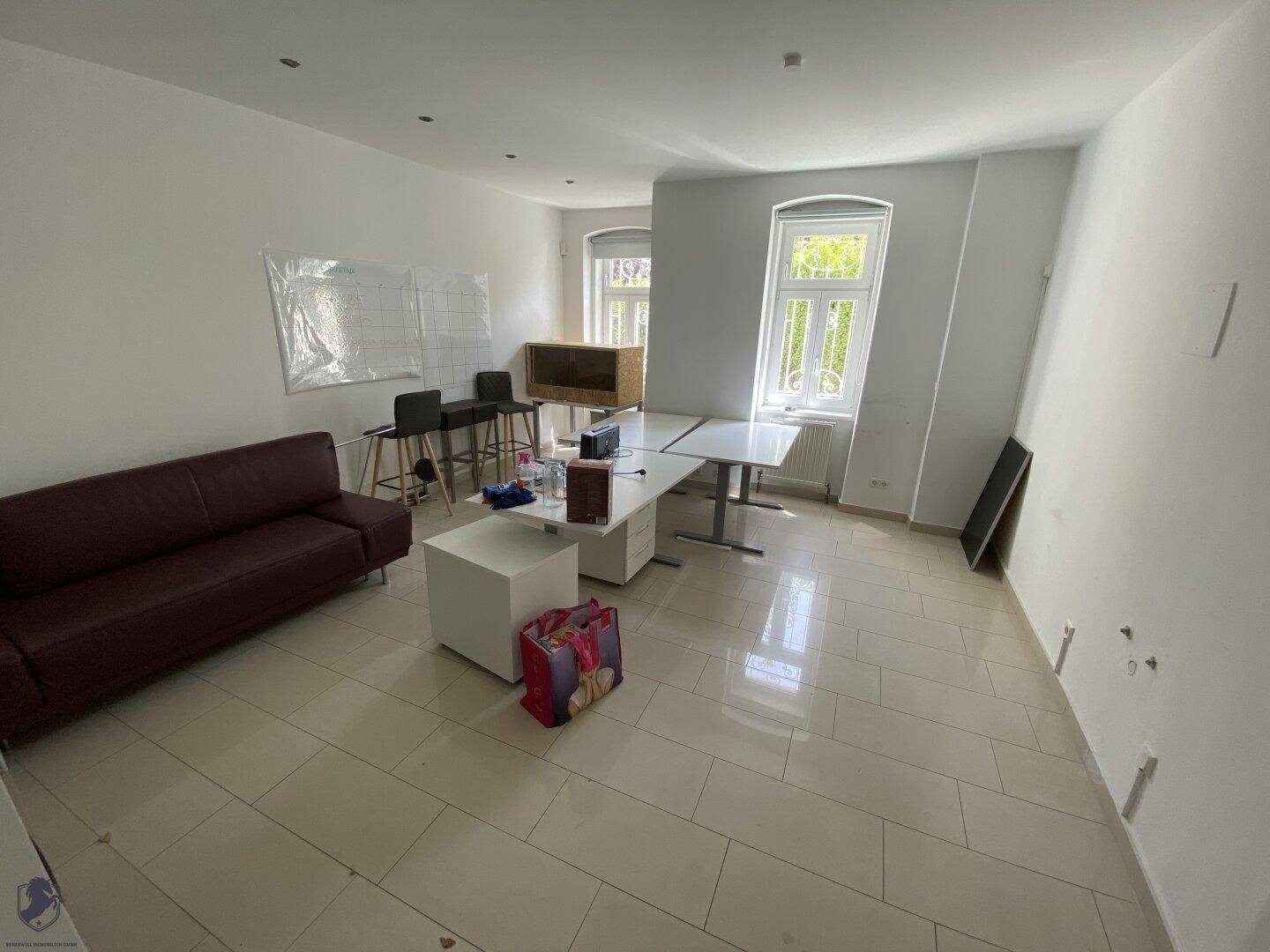 Geräumiger Raum mit Fliesenboden, Sofa und mehreren Schreibtischen, ideal als Büro.
