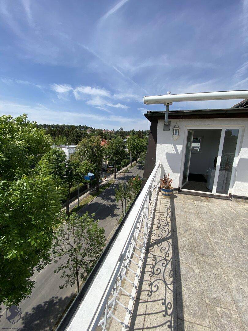Dachterrasse mit Fliesenboden, schmiedeeisernem Geländer und Blick auf die Straße und Bäume.