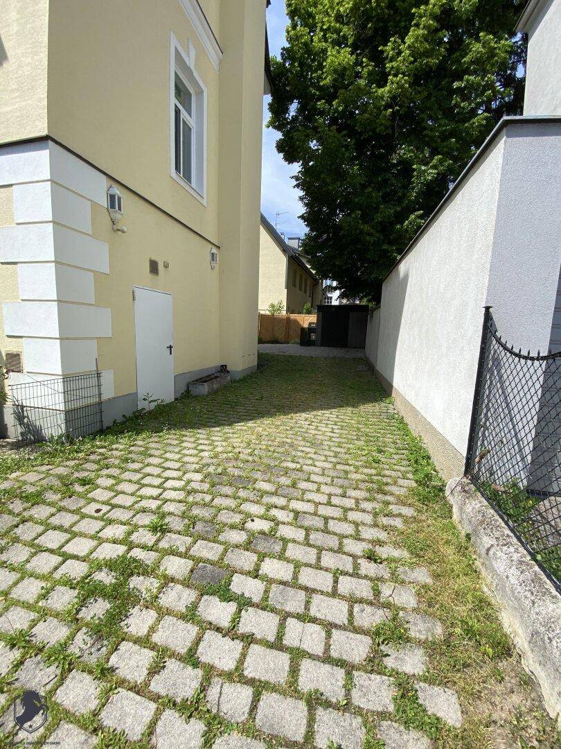 Gepflasterter Weg neben dem Gebäude, führt zu einem hinteren Bereich mit weiteren Bäumen.