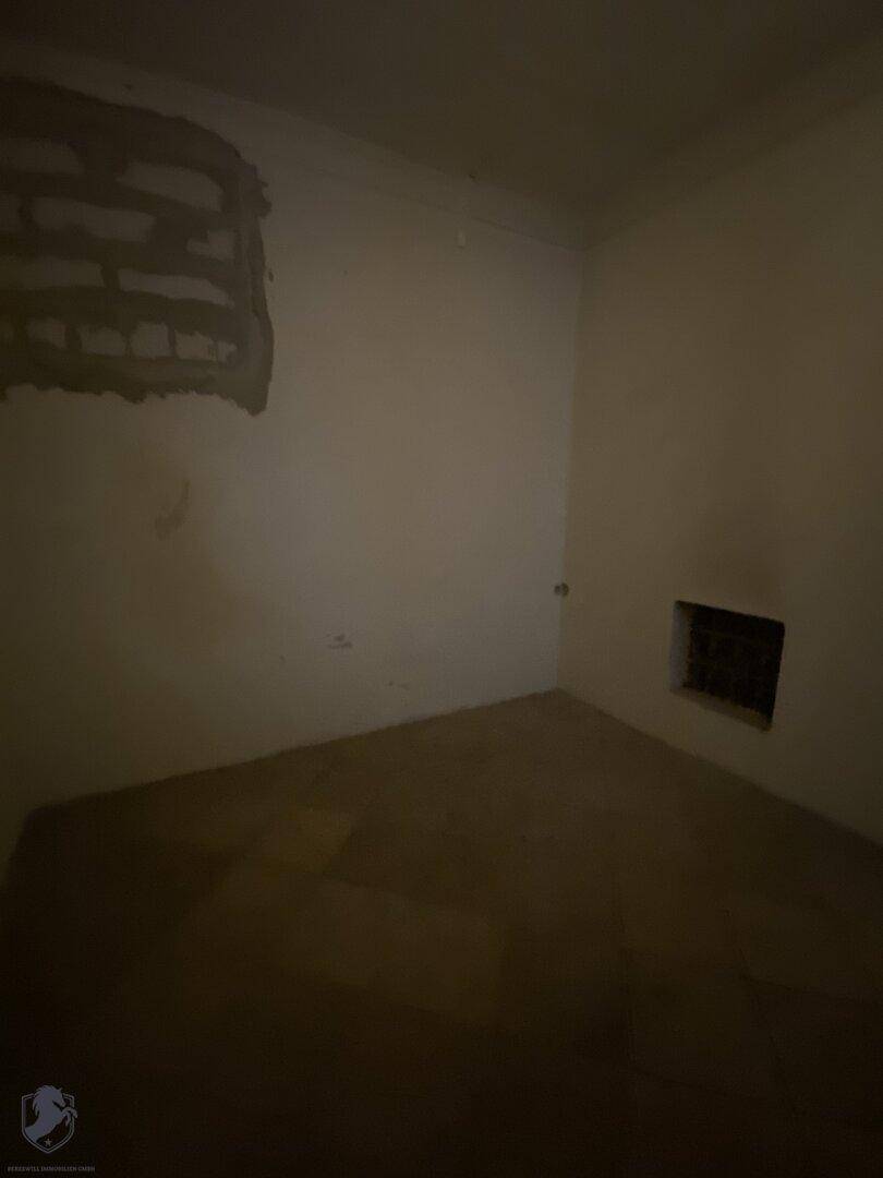 Ein dunkler, leerer Raum mit einfachen Wänden und einem kleinen Fenster, das wenig Licht hereinlässt.