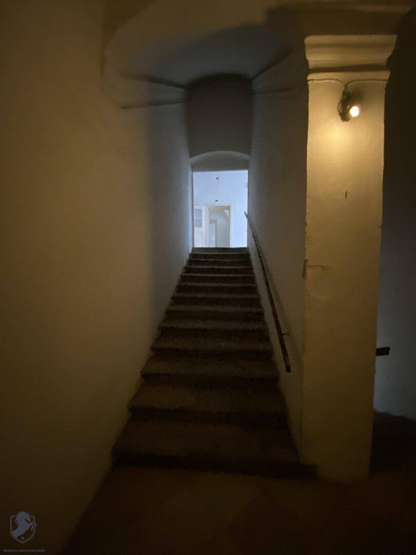 Historisches Treppenhaus mit Steinstufen und einem Handlauf, beleuchtet von einer Wandleuchte.