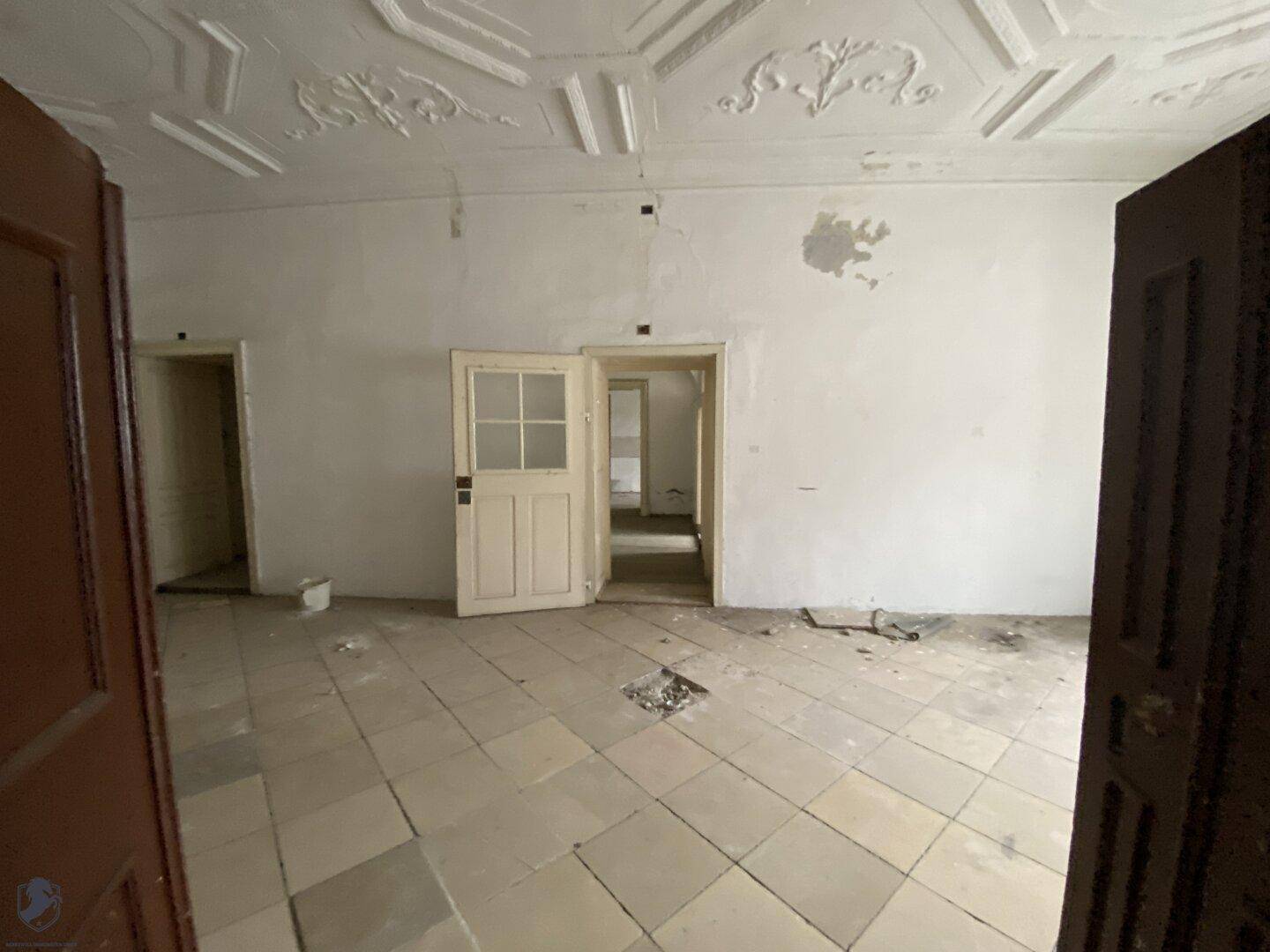 Geräumiger, unrenovierter Raum mit dekorativer Stuckdecke und Fliesenboden, bereit zur Sanierung.