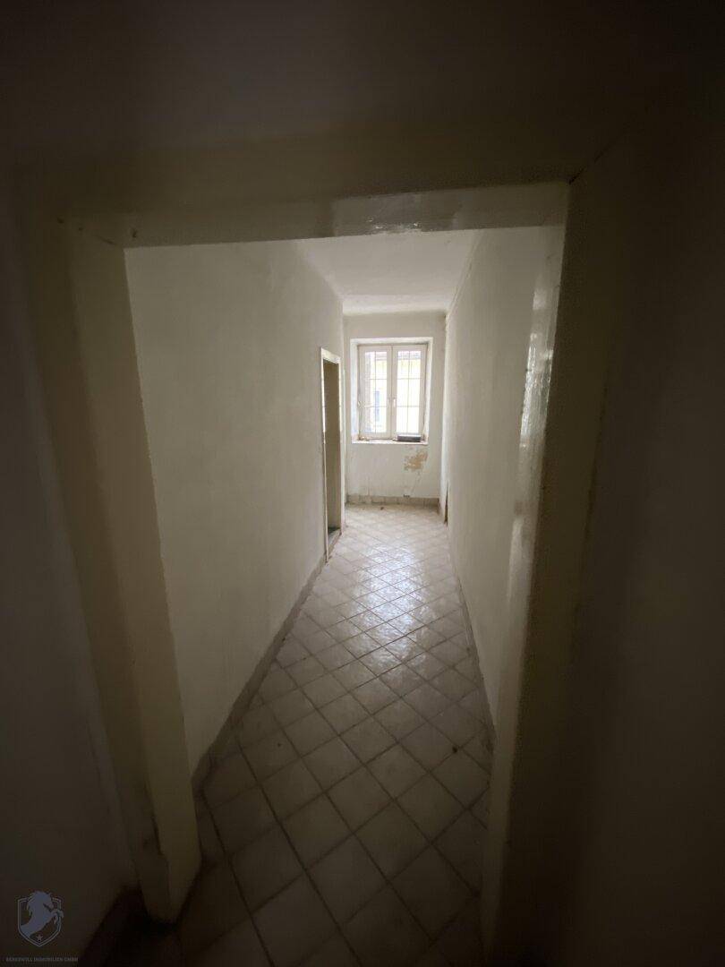 Langer, schmaler Flur mit Fliesenboden und einem Fenster am Ende, das Tageslicht hereinlässt.