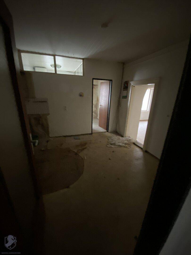 Dunkler, unrenovierter Raum mit mehreren Türen und einem Fensterband oben, das Licht spendet.