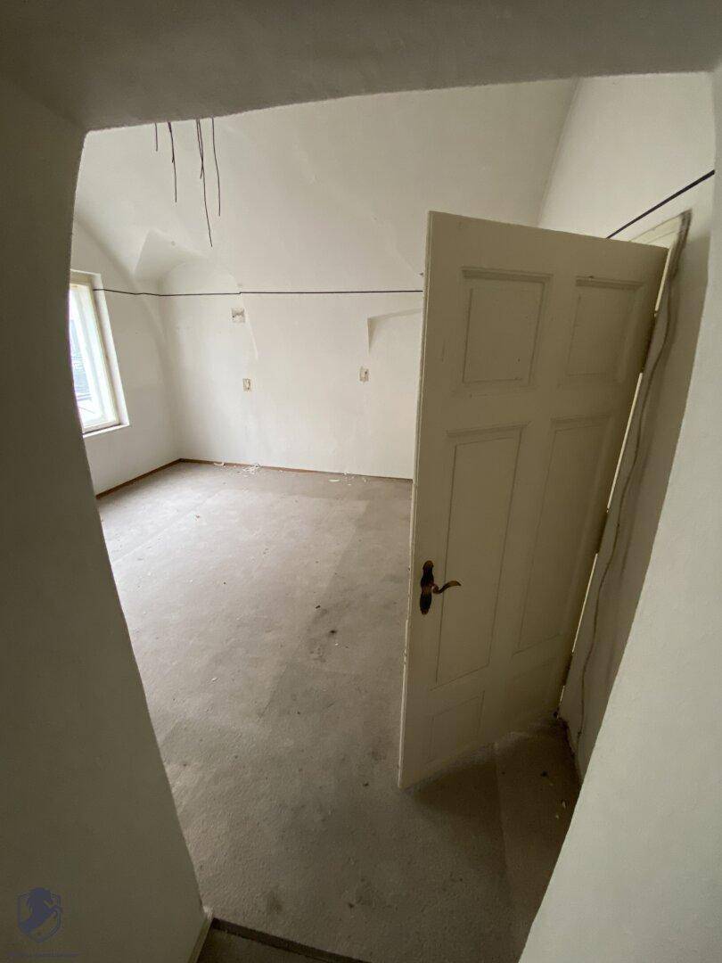 Heller, unrenovierter Raum mit schrägen Wänden und einem Fenster, das Tageslicht hereinlässt.