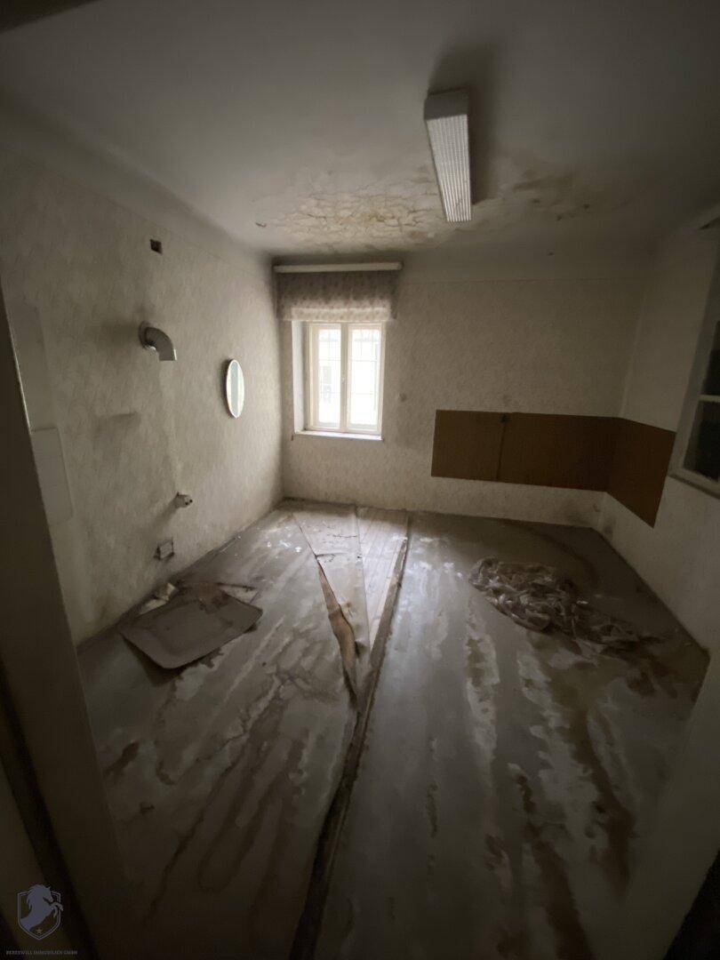 Dunkler, unrenovierter Raum mit stark beschädigtem Boden und einem kleinen Fenster, das wenig Licht spendet.