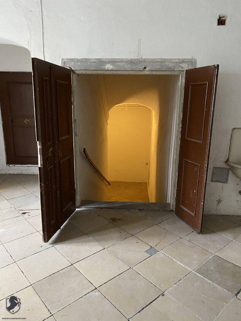 Offene Doppeltür führt zu einem dunklen Treppenhaus mit Steinstufen, das nach unten führt.