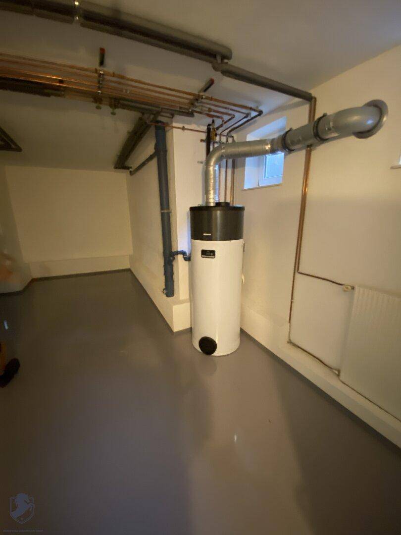 Hauswirtschaftsraum im Keller mit Warmwasserspeicher, sichtbaren Rohren und kleinem Fenster.