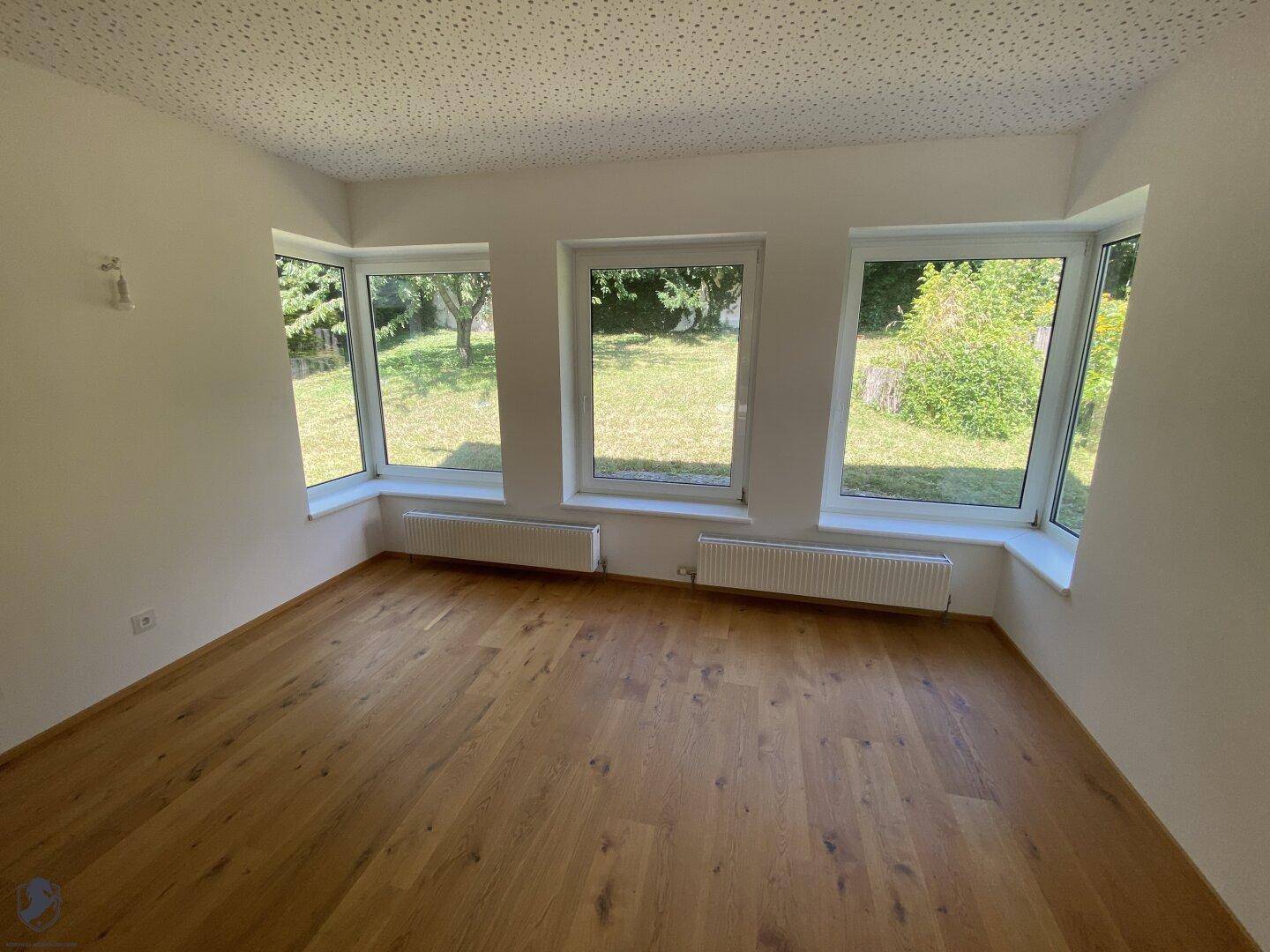 Heller Raum mit Holzdielenboden, weißen Wänden und Erkerfenstern mit Gartenblick.