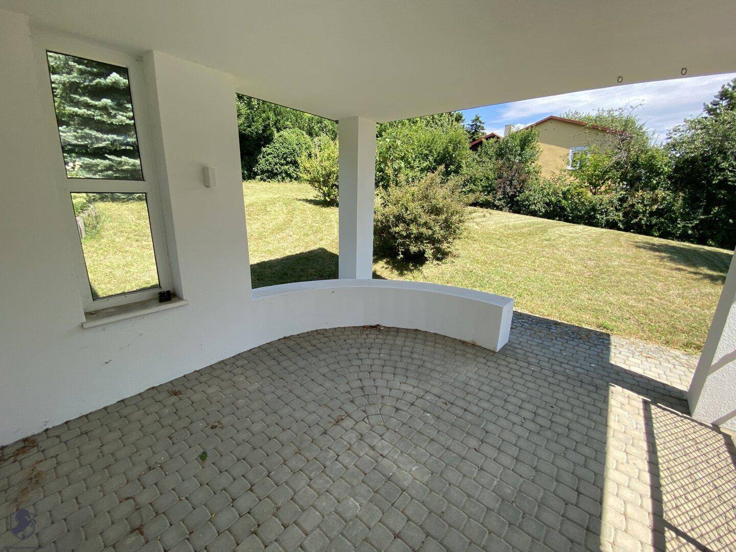 Überdachter Terrassenbereich mit gemustertem Pflaster und geschwungener Mauer, mit Blick auf den Rasen.