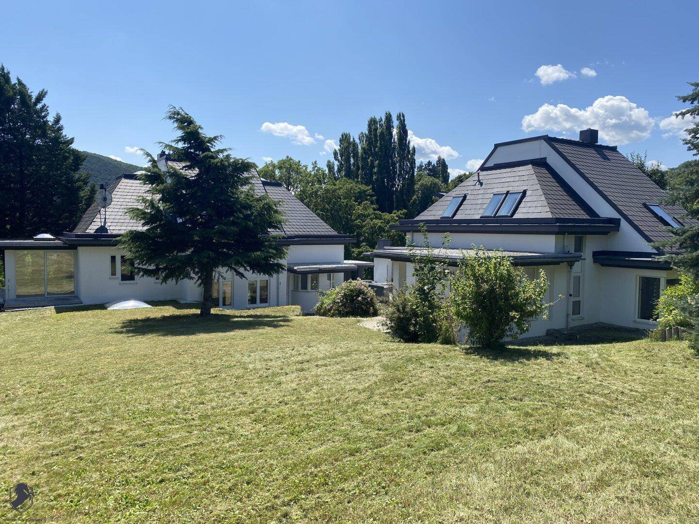 Zwei moderne Häuser mit dunklen Dächern und weißer Fassade, umgeben von einem weitläufigen Rasen und Bäumen.