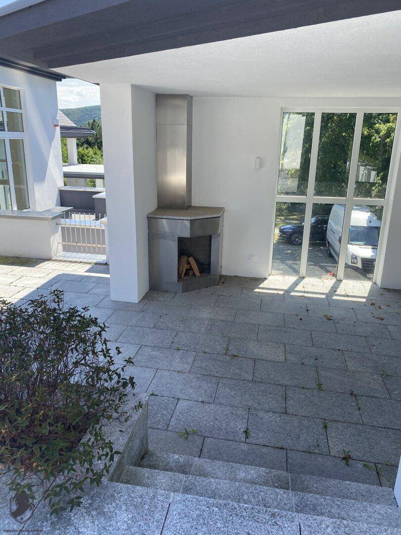 Überdachter Terrassenbereich mit Steinfliesen, Außenkamin und großen Glasfenstern zum Innenbereich.
