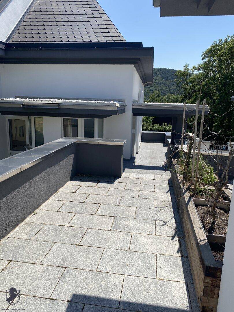 Weitläufige gepflasterte Terrasse mit niedriger Mauer und Blick auf die moderne Architektur des Hauses.