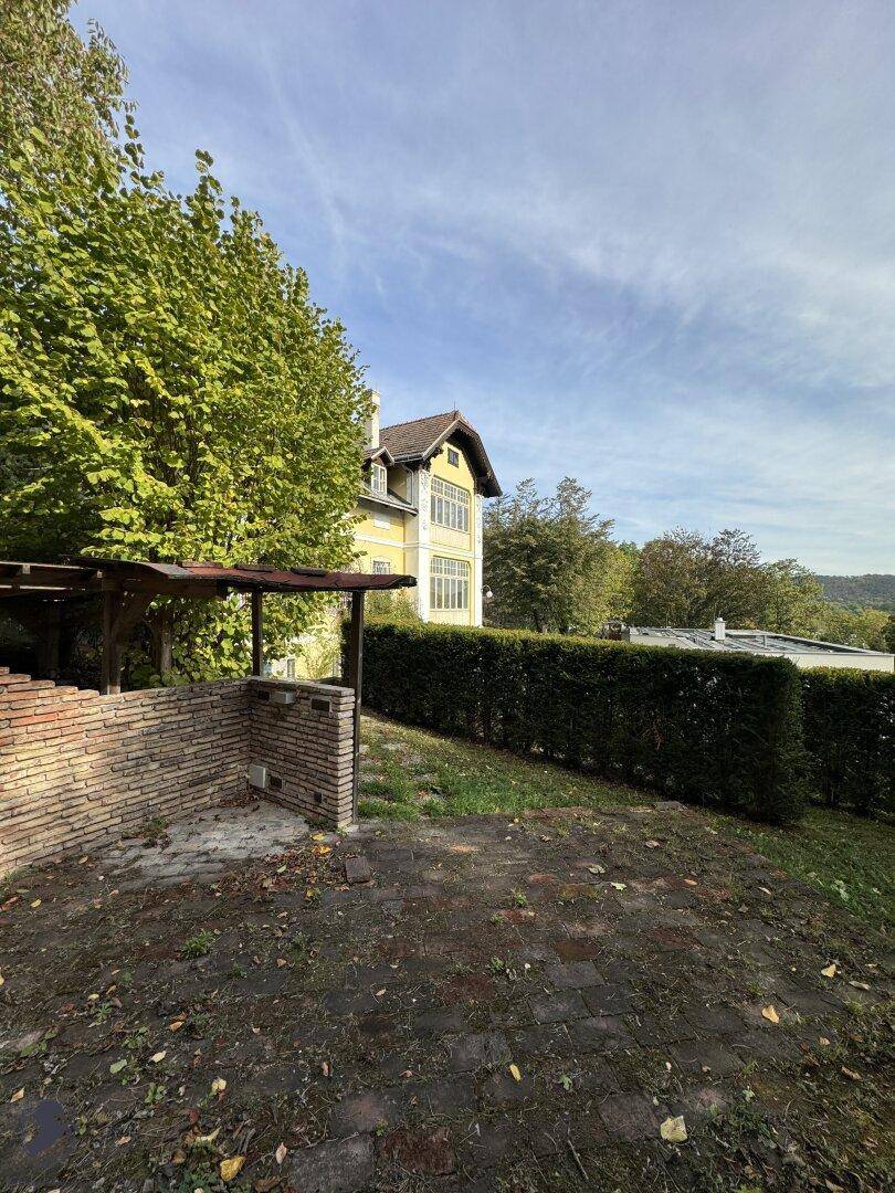 Gartenbereich mit einer gemauerten Grillstelle und Hecke, die zur historischen Villa führt.
