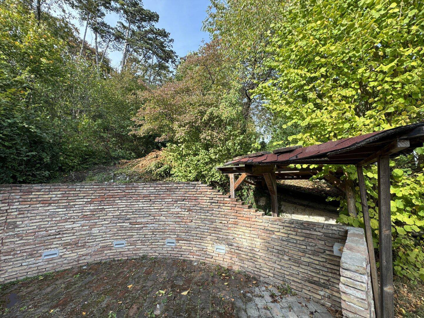 Gemauerte Struktur mit Holzdach in einem bewaldeten Gartenbereich, umgeben von Bäumen.