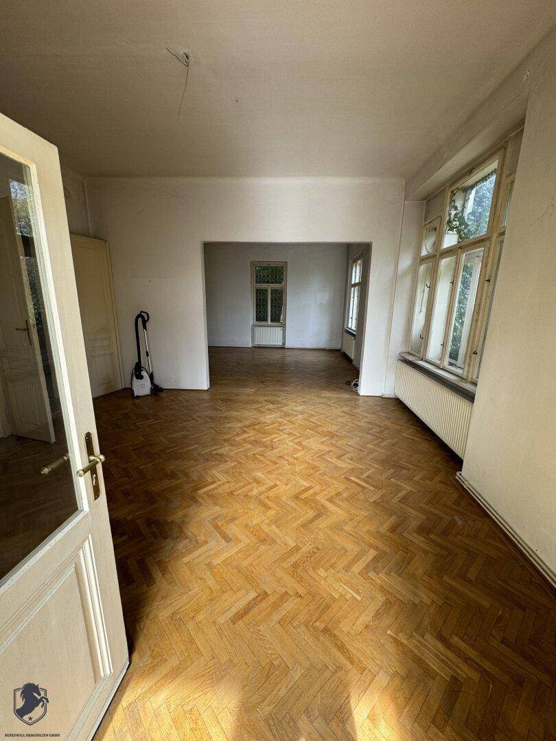 Geräumiger Wohnbereich mit Fischgrätparkett und hohen Decken, der in weitere Räume führt.