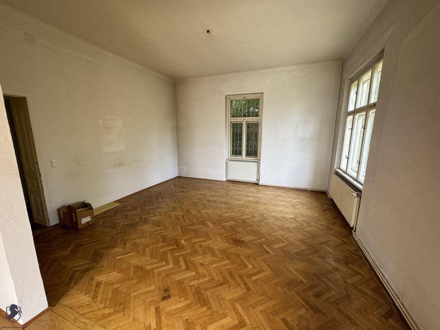 Helles Zimmer mit Fischgrätparkett und einem großen Fenster, das viel Tageslicht hereinlässt.