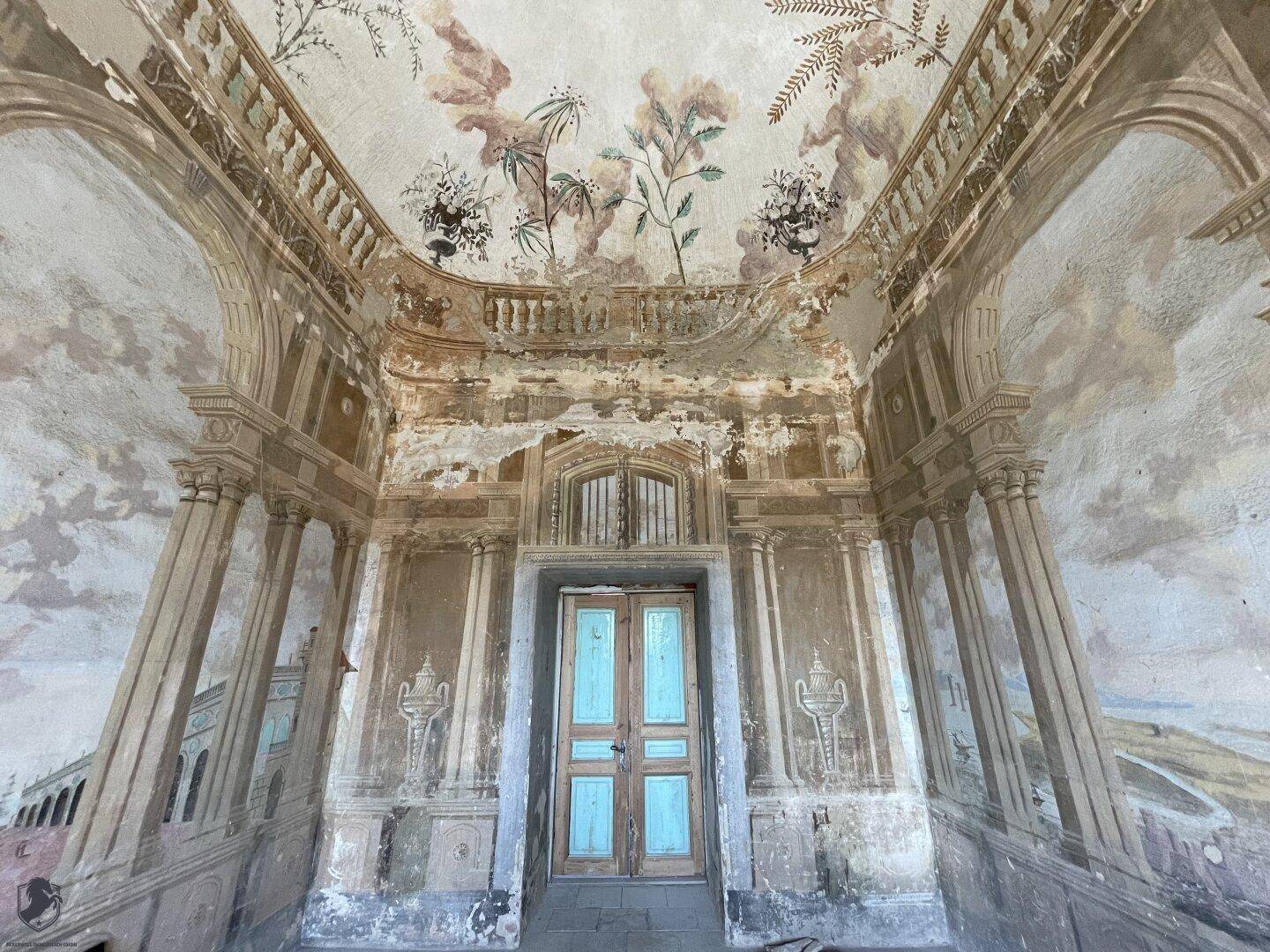 Historischer Raum mit verblassten Wand- und Deckenmalereien sowie einer alten Holztür.