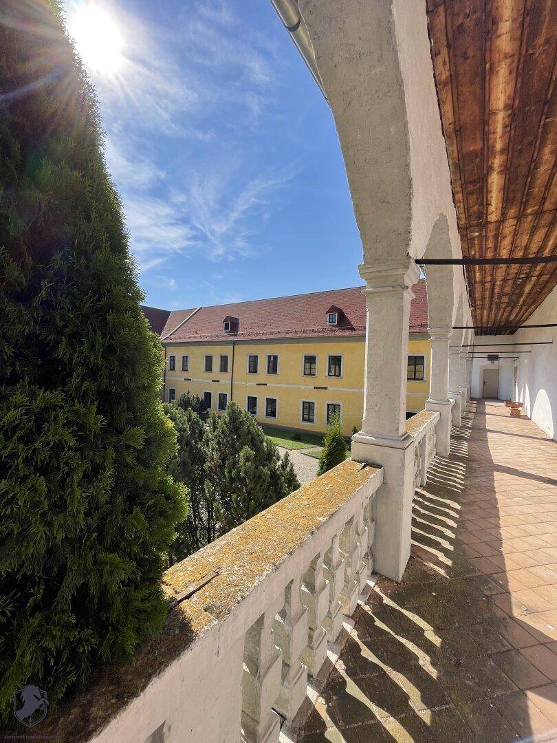 Überdachter Arkadengang mit Balustrade und Blick auf die sonnige Schlossfassade und grüne Bäume.