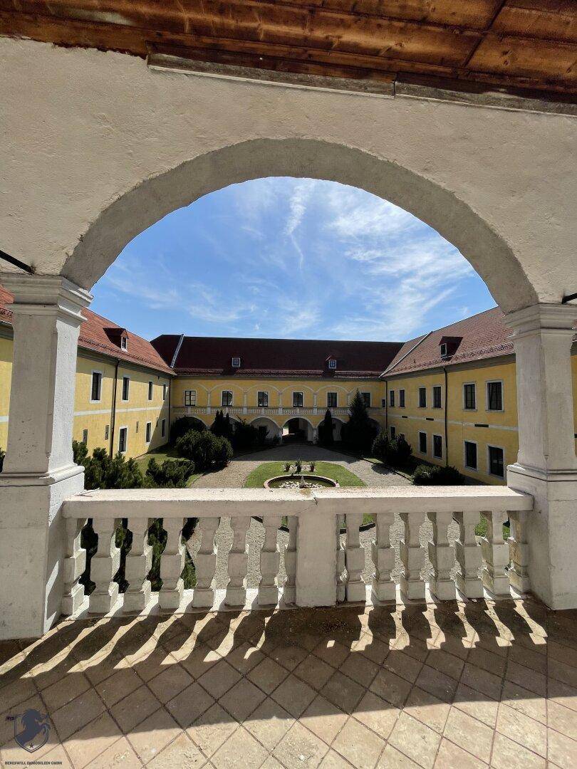 Blick durch einen Arkadenbogen auf den sonnigen Innenhof mit Brunnen und historischem Schlossgebäude.