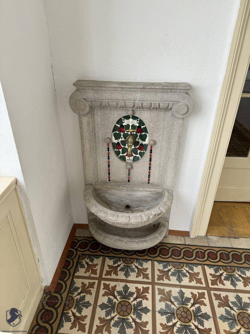 Eingangsbereich mit dekorativen Fliesen und einem antiken Wandbrunnen als Blickfang.