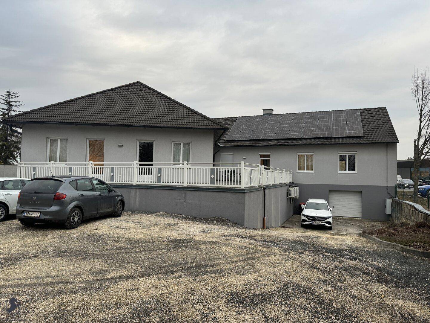 Rückseite des Gebäudes mit einer großen Terrasse, Solaranlage auf dem Dach und weiteren Parkmöglichkeiten.