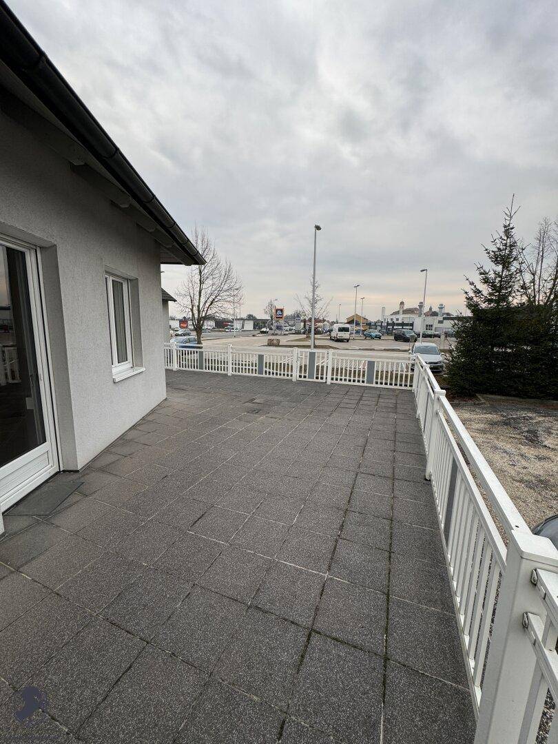 Gepflasterte Terrasse mit weißem Geländer, die einen Blick auf den Parkplatz und die Umgebung bietet.