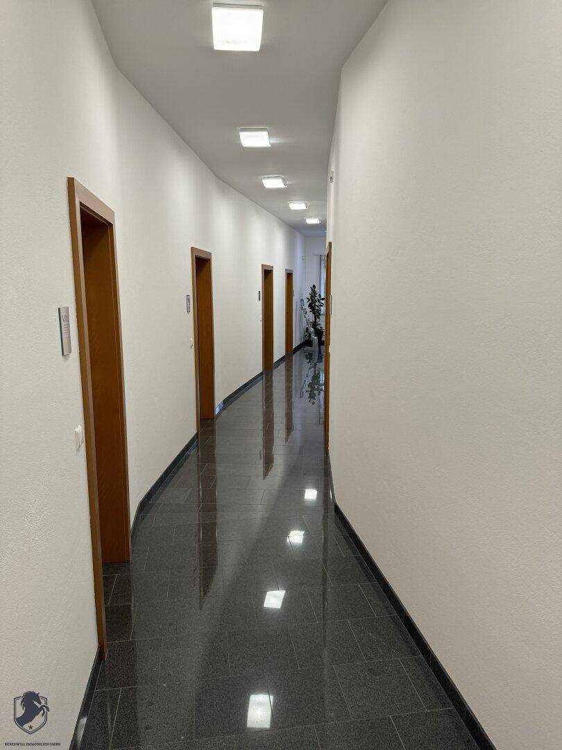 Langer, heller Flur mit dunklem Fliesenboden und mehreren Holztüren, die zu den Büroräumen führen.