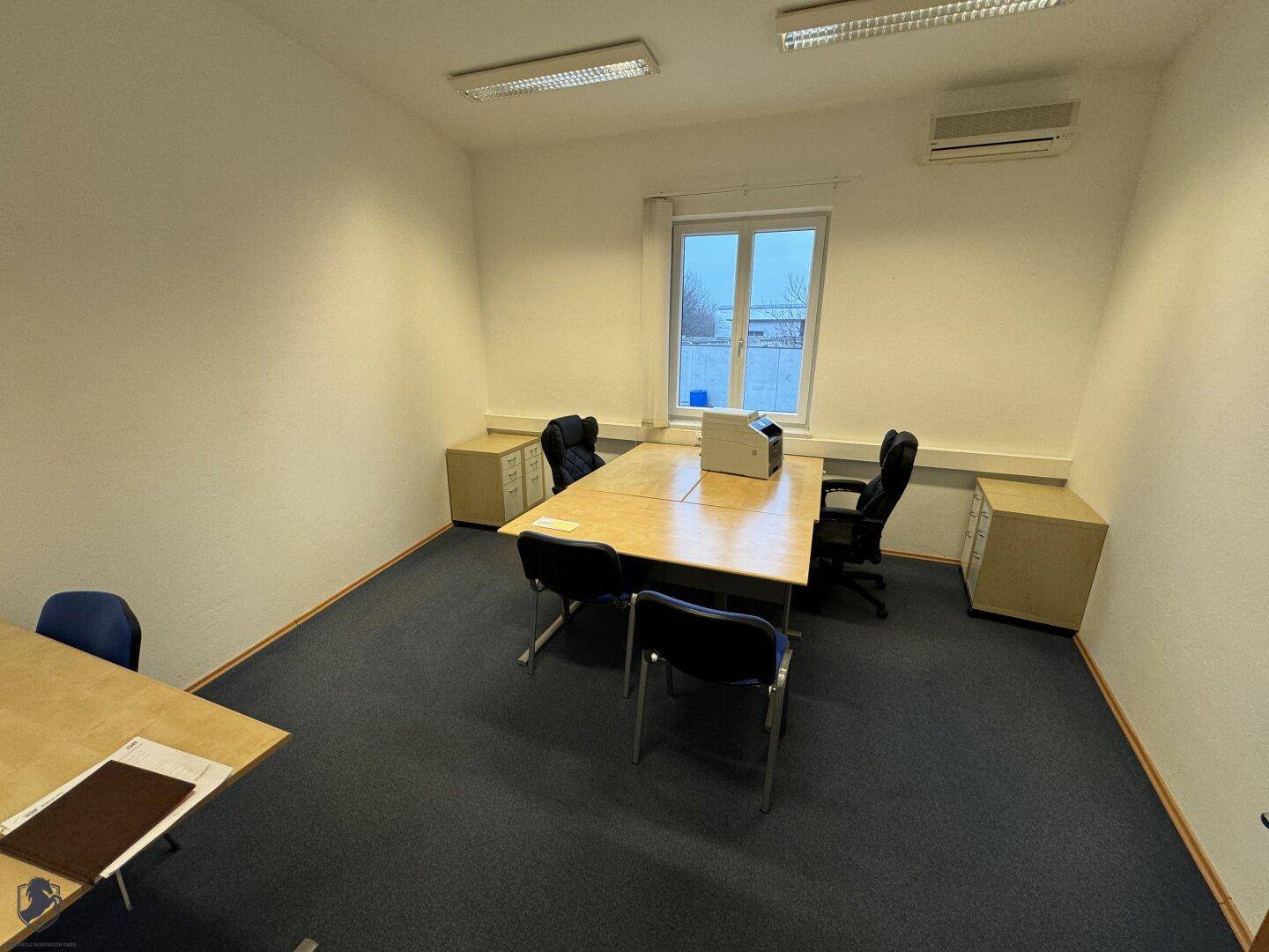 Funktionales Büro mit einem Besprechungstisch, Bürostühlen und einem Fenster, das Tageslicht hereinlässt.