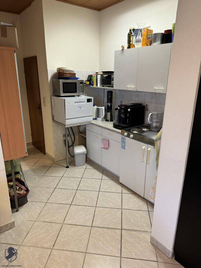 Kleine Küchenzeile mit weißen Schränken, Mikrowelle, Kaffeemaschine und Fliesenboden.