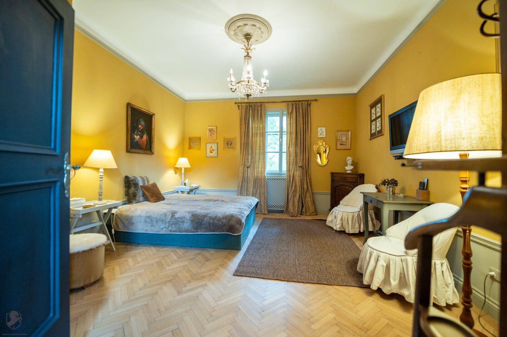 Gemütliches Schlafzimmer mit gelben Wänden, Kronleuchter und klassischem Fischgrätparkettboden.