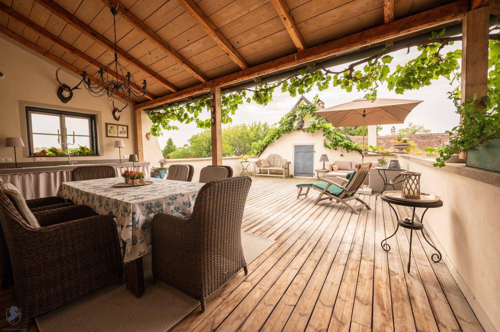 Überdachte Holzterrasse mit Esstisch, Liegestühlen und üppiger Begrünung.