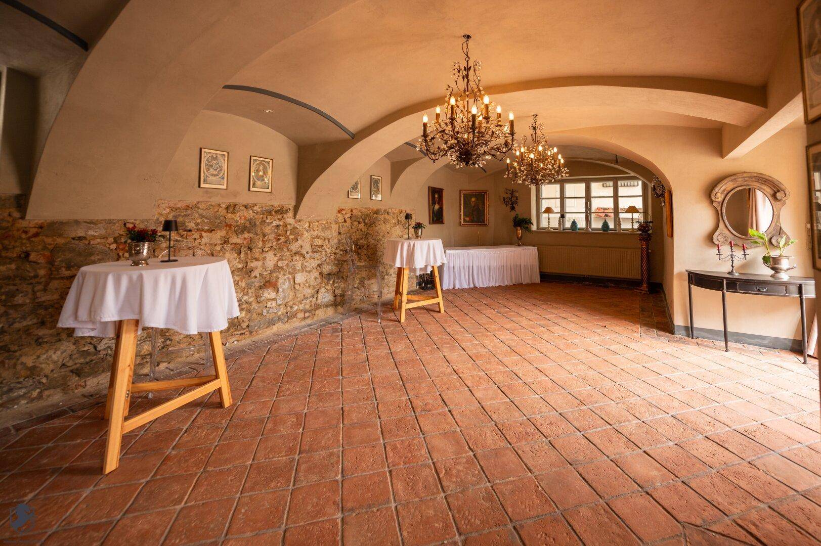 Historischer Gewölberaum mit Terrakottaboden, freigelegter Steinwand und mehreren Kronleuchtern.