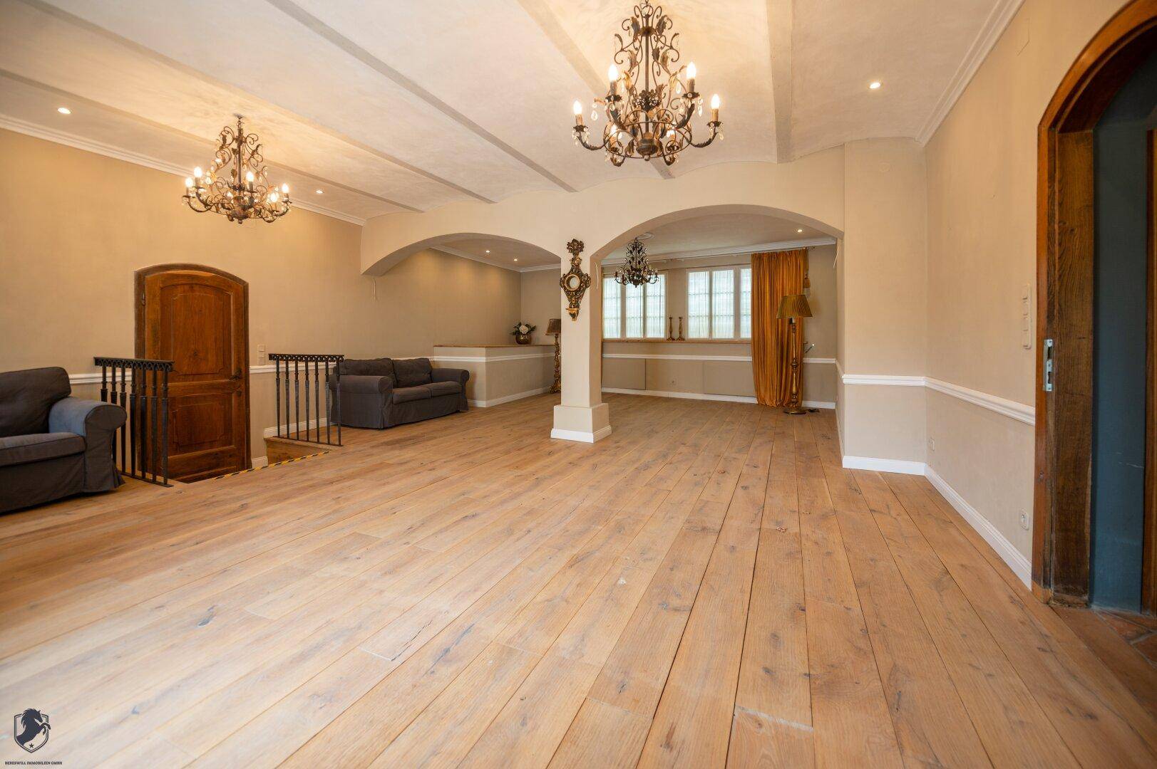 Großer offener Raum mit breiten Holzplanken, eleganten Bögen und zwei Kronleuchtern.