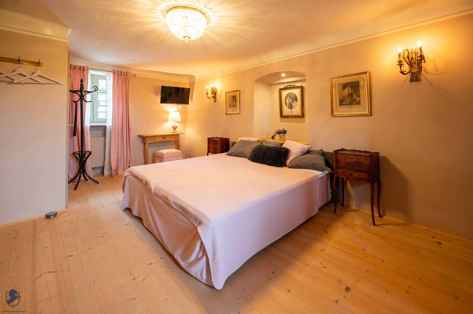 Schlafzimmer mit großem Bett, rosa Tagesdecke und hellem Holzplankenboden.