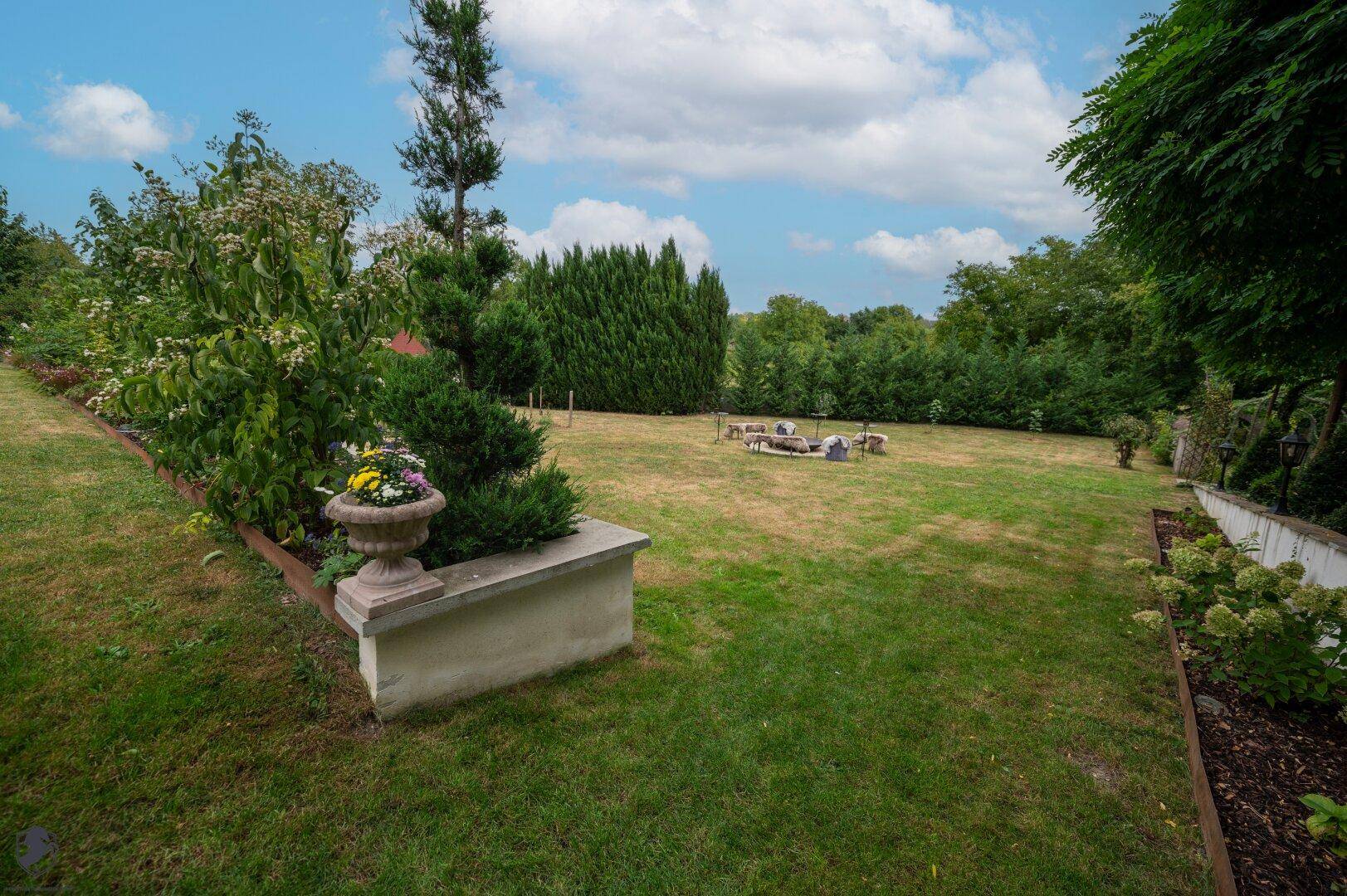 Weitläufige Rasenfläche im Garten mit Feuerstelle und umgebenden Bäumen.
