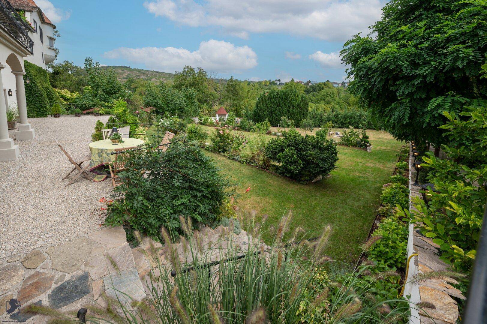 Erhöhter Blick auf einen gepflegten Garten mit Rasen, Blumenbeeten und Sitzgelegenheiten.