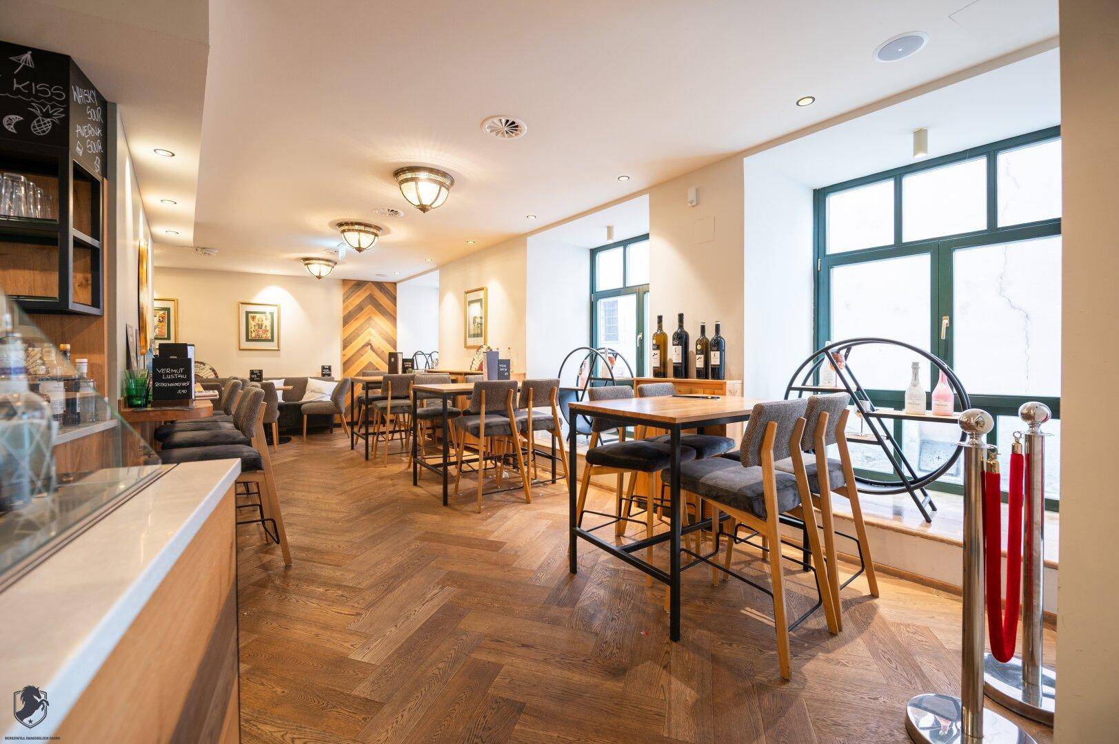 Heller Restaurantbereich mit Holzboden, großen Fenstern und verschiedenen Sitzgelegenheiten.