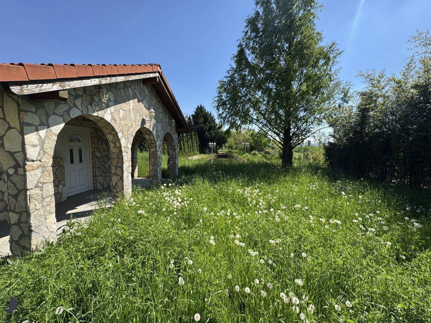 Rustikales Steingebäude mit Rundbögen und einem weitläufigen, blühenden Garten im Vordergrund.