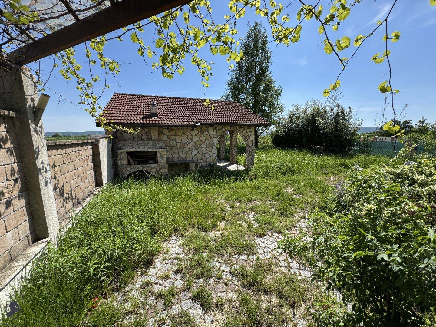 Außenküche mit Steingrill und gepflastertem Bereich, umgeben von einem grünen Garten.