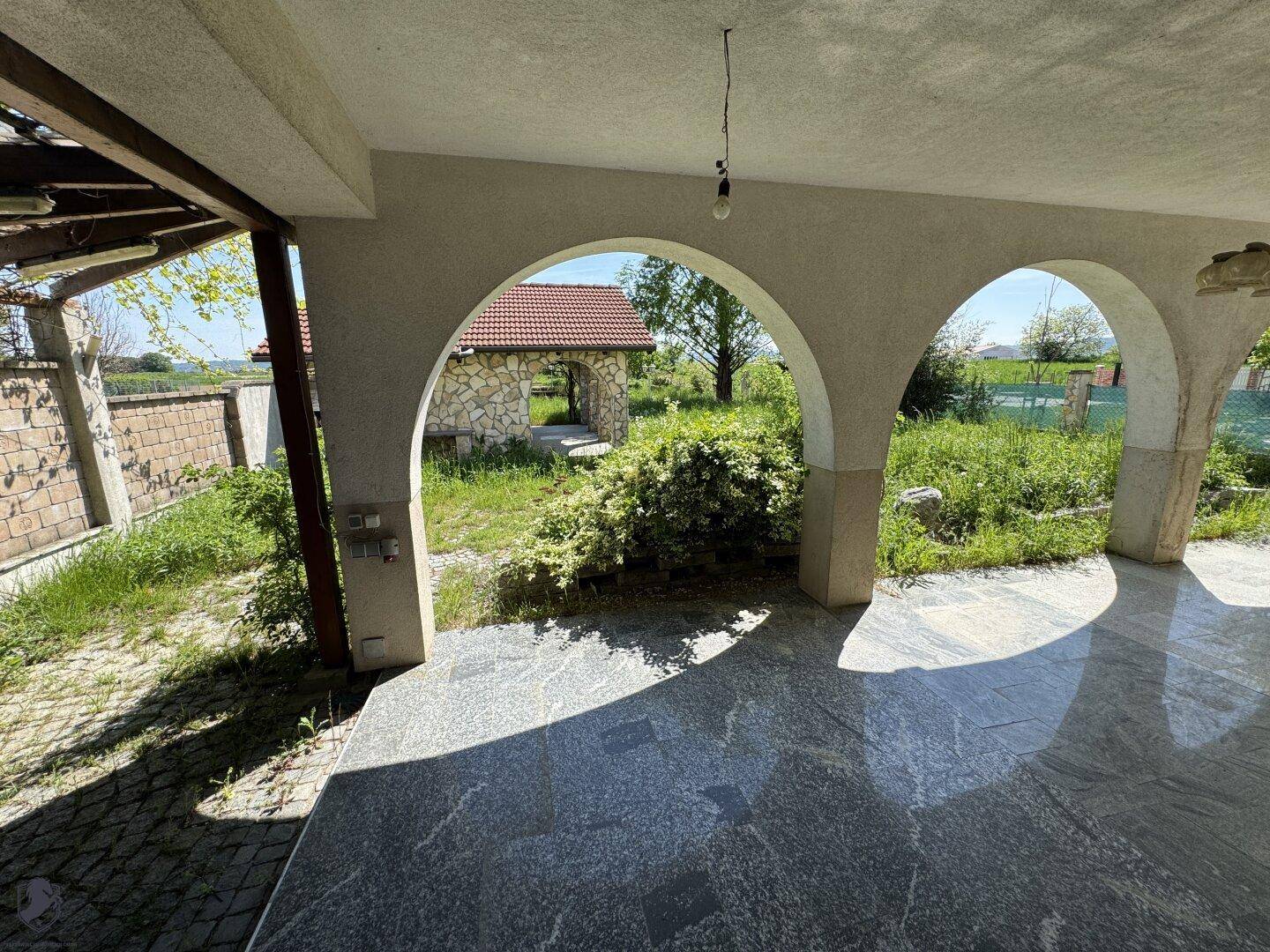 Überdachte Terrasse mit Fliesenboden und Rundbögen, bietet einen Blick in den grünen Garten.