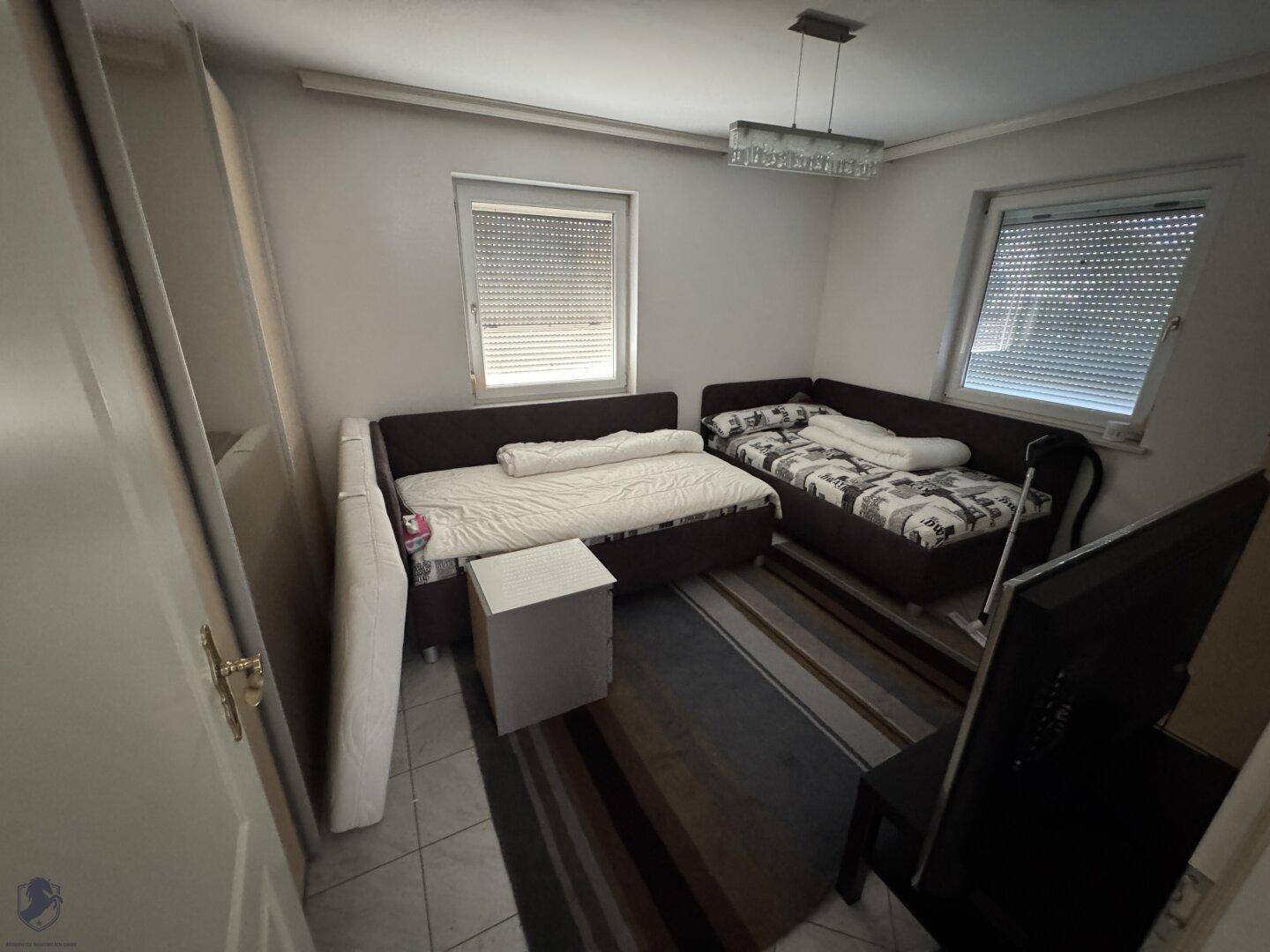 Schlafzimmer mit zwei Betten, kleinem Tisch und zwei Fenstern mit Jalousien.