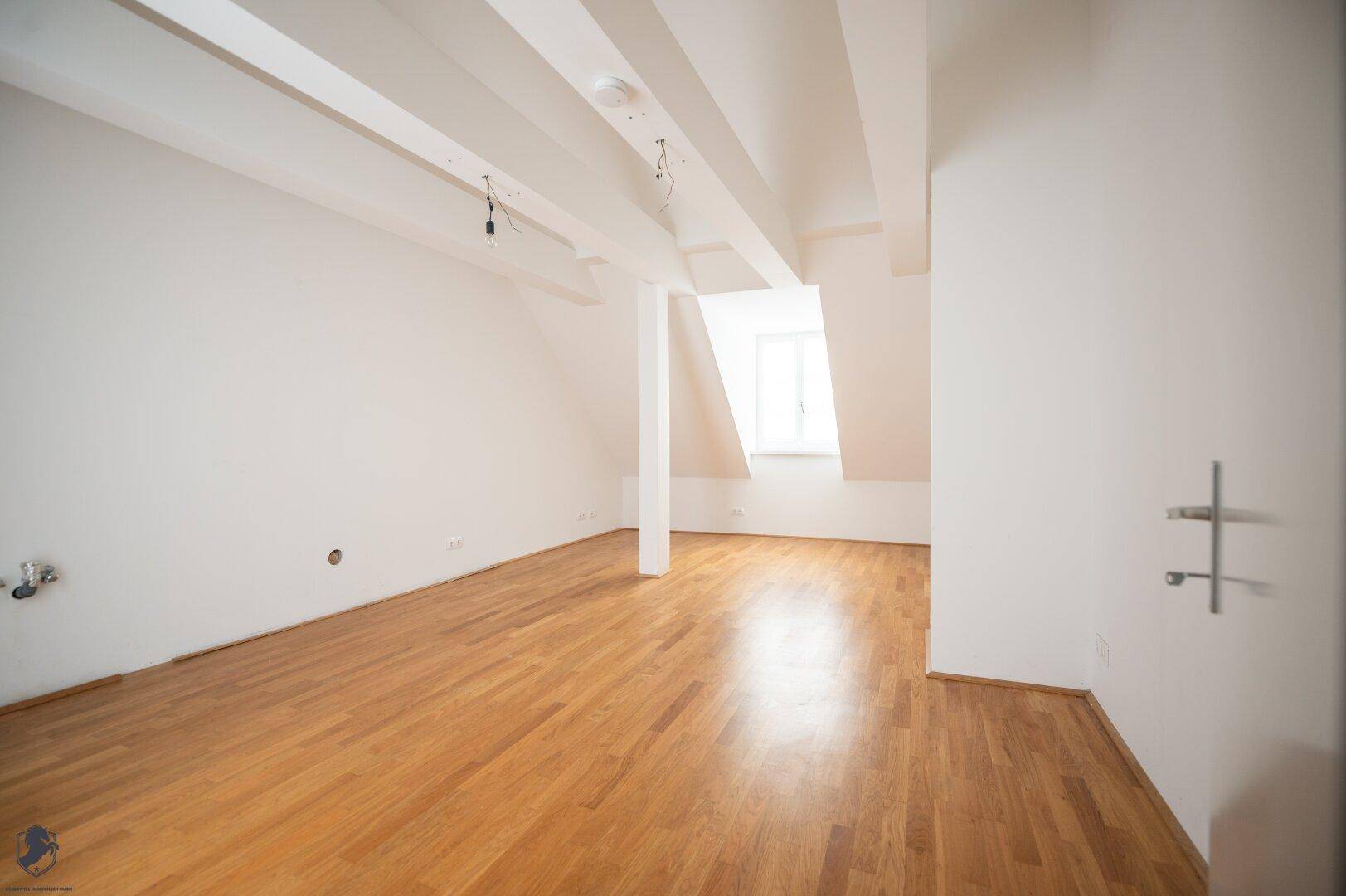 Geräumiger, leerer Raum mit Holzboden, weißen Wänden und sichtbaren Deckenbalken im Dachgeschoss.