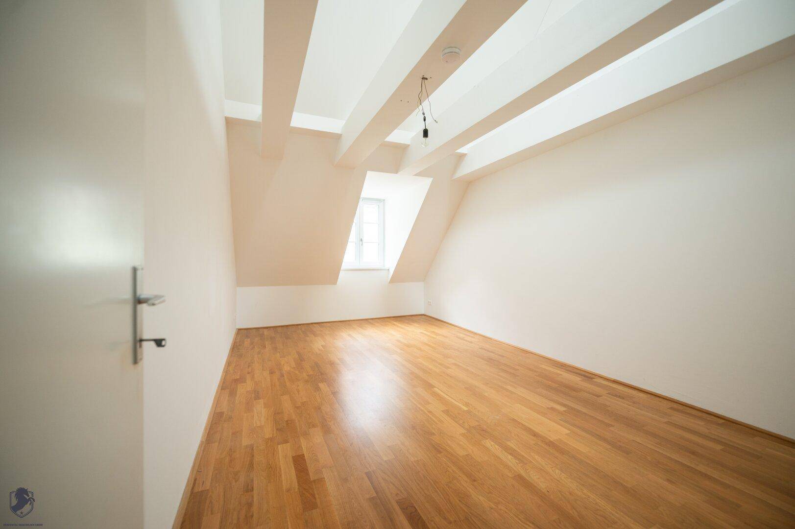 Heller Raum mit Holzboden, weißen Wänden und sichtbaren Deckenbalken unter dem Dach.
