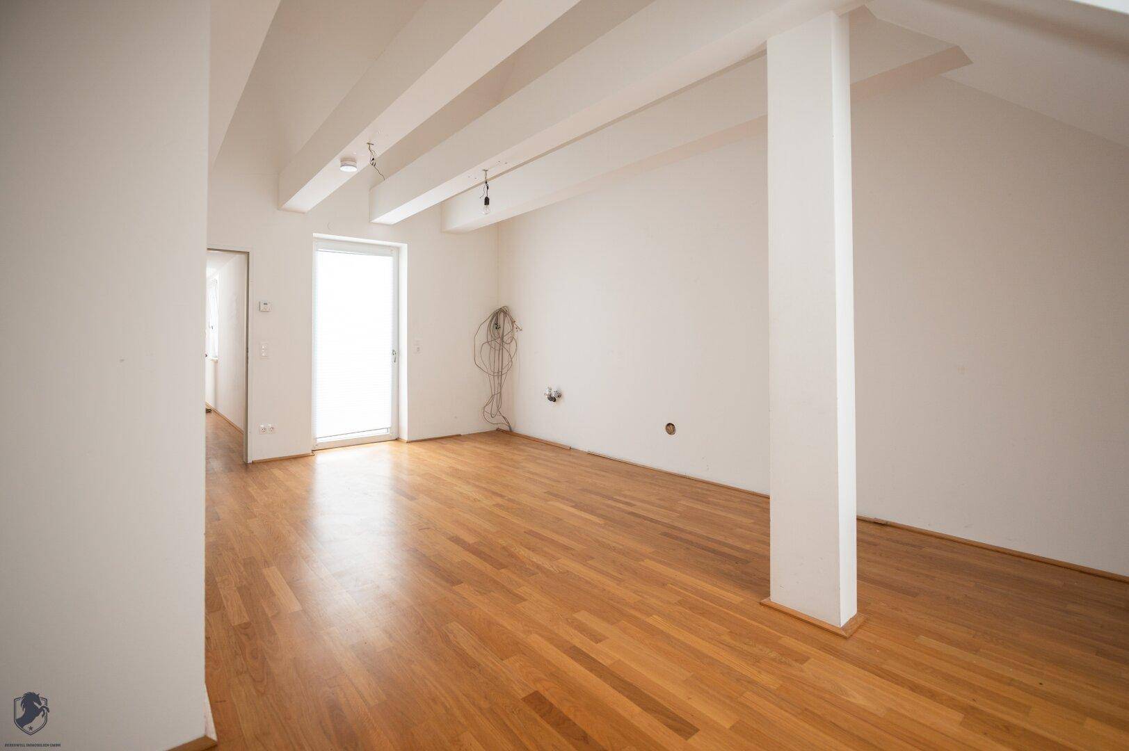 Geräumiger Raum mit Holzboden, weißen Wänden und Zugang zu einem Außenbereich.