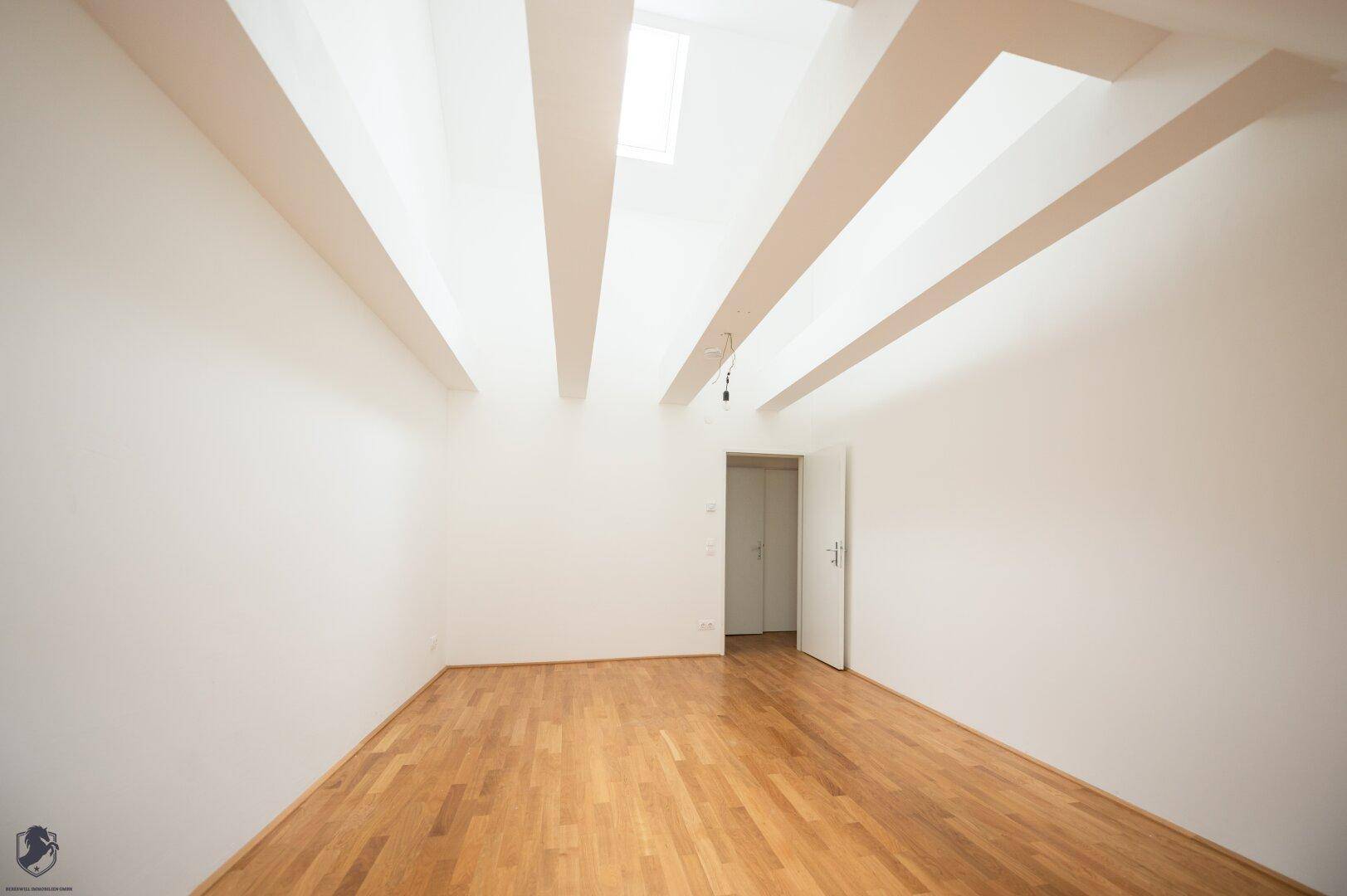 Heller Raum mit Holzboden, weißen Wänden, sichtbaren Deckenbalken und einem Dachfenster.